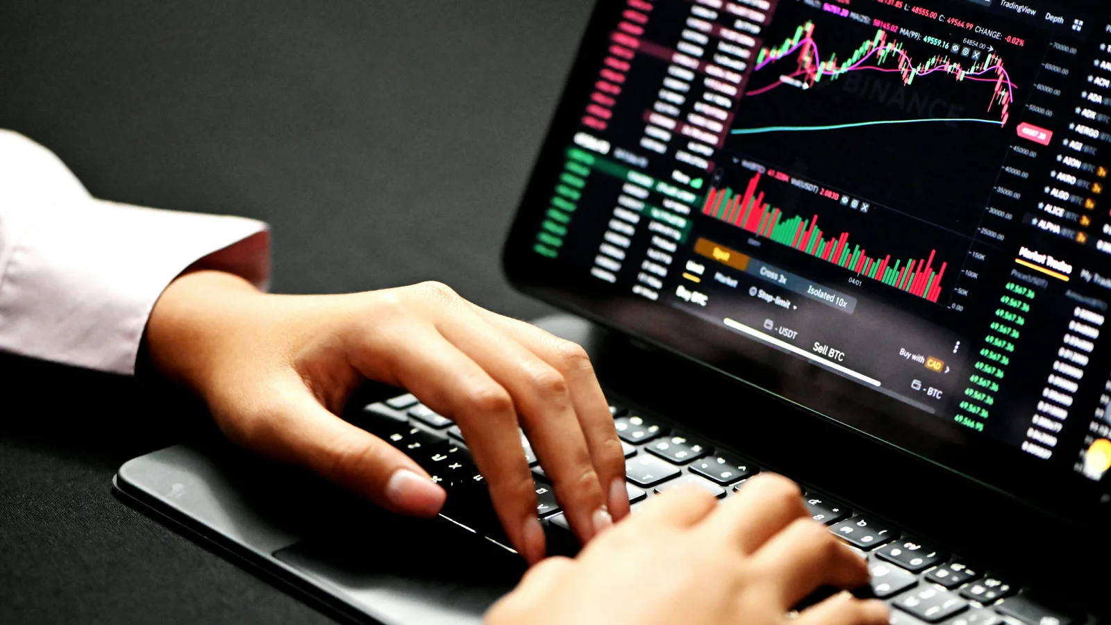 Manos de un trader tecleando en un ordenador portátil mientras analiza un gráfico de cotizaciones y volúmenes de criptomonedas en la pantalla.