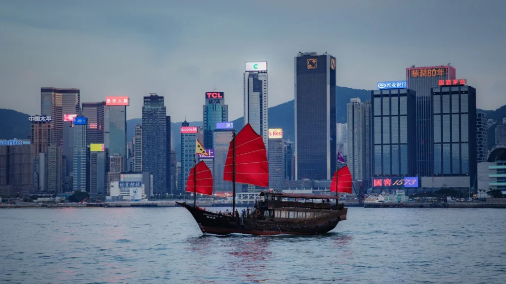 Barco tradicional con velas rojas navegando por Victoria Harbour frente al skyline financiero de Hong Kong.